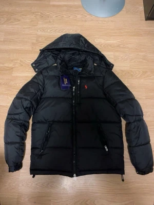 Svart pufferjacka från Polo Ralph Lauren - Svart pufferjacka från Polo Ralph Lauren med avtagbar huva, dragkedjor på fickor och bröstficka samt klassisk röd logga på bröstet. Jackan är vadderad och har en sportig look, perfekt för kalla dagar. Materialet är slitstarkt och håller värmen bra.