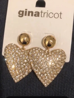 Glittriga hjärtörhängen Gina Tricot - Säljer ett par stora, glittriga örhängen från Gina Tricot i form av hjärtan. Örhängena är guldfärgade och täckta med gnistrande stenar som ger en lyxig känsla. Perfekta för dig som vill sticka ut och addera extra bling till din look.