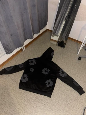 Denim tears hoodie - Säljer en svart hoodie med gråa blommönster över hela tröjan. Hoodien har en klassisk huva och ribbade muddar vid ärmslut och nederkant. Perfekt för dig som gillar streetwear och vill sticka ut lite extra.