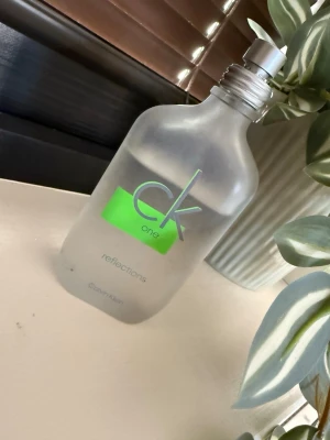 CK One Reflections parfymflaska - Säljer en CK One Reflections parfym från Calvin Klein. Flaskan är frostad och har en stilren, rektangulär form med rundade kanter. Märket och namnet syns i grönt och silver på framsidan. Flaskan är i glas och har en metallfärgad kork. (Skulle säga att det är runt 70ml kvar) (Flaskan har några få fläckar som jag inte fått bort, men det går säkert på annat sätt)