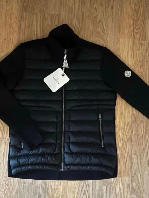 Moncler Cardigan - Snygg svart dunjacka från Moncler med quiltad design och två dragkedjeförsedda fickor framtill. Jackan har hög krage och ikonisk Moncler-logga på ärmen. Materialet är en kombination av dunfyllning och mjuka tygpartier på ärmarna för extra komfort och stil.