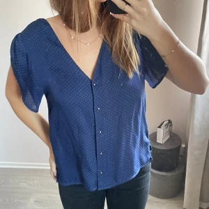 Blå prickig blus med v-ringning - Söt blå blus med små prickar och v-ringning. Blusen har korta puffärmar och knappar framtill. Storlek S. Inga defekter, perfekt inför sommaren. 
