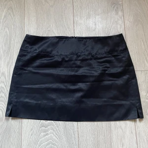 Svart satin kortkjol - Snygg svart kortkjol i satin med diskret glansig finish. Kjolen har en enkel, rak passform och liten slits på sidorna. Från märket French connection i storlek 6, ca xs. Hör av er vid frågor om mått osv💗