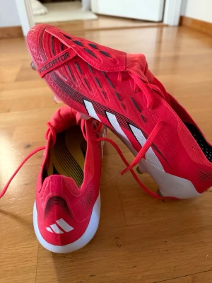 PREDATOR ELITE FOLD-OVER TONGUE FIRM GROUND FOTBOLLSSKOR - Säljer ett par röda Adidas Predator fotbollsskor med vita och svarta detaljer. Skorna har snörning, klassisk låg profil och slitstark syntetisk ovandel. Ikoniska tre ränder på sidan och texten Predator på tån. Säljer dessa då jag ej har användning från dessa då jag har ett annat par. Säg gärna till om fler bilder!