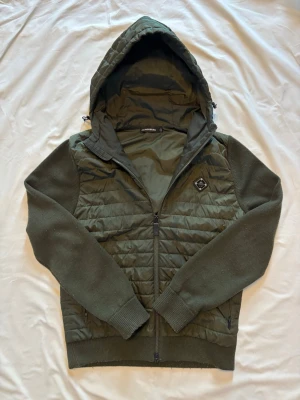 Mörkgrön jacka med huva från J.Lindberg - Snygg mörkgrön jacka från J.Lindberg med quiltad front och rygg, stickade ärmar och muddar. Jackan har dragkedja, två sidofickor med dragkedja och en stor huva. Perfekt för dig som vill ha en casual och sportig look. OBS den är lagad på högra ärmen