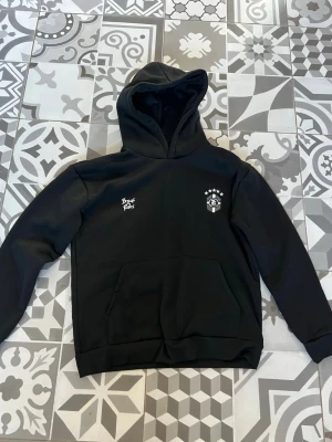 Svart hoodie från Bror Pals - Svart hoodie från Bror Pals med tryck på bröstet och stor ficka framtill. Klassisk huva och ribbade muddar vid ärmar och nederkant. Perfekt för en chill och avslappnad stil. Och det är  Asian S, storlek