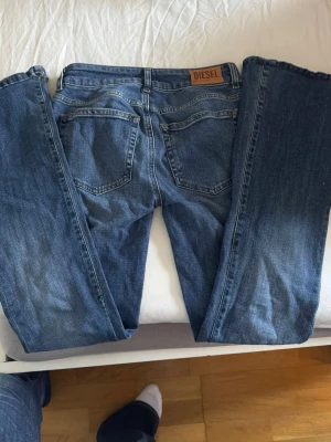 Blå raka jeans från Diesel - Snygga blå lågmidjade jeans från Diesel med klassisk femficksmodell och raka ben. Jeansen har en mörk tvätt och detaljer som läderpatch med logga bak i linningen. Tillverkade i slitstarkt denimtyg, perfekta för en avslappnad och trendig stil.
