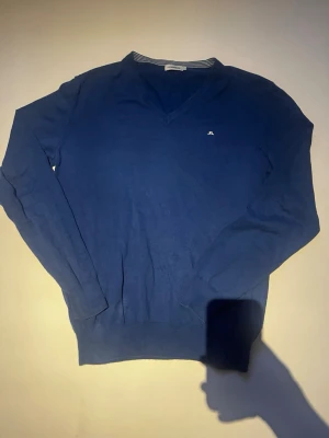 Blå V-ringad tröja J.Lindeberg - Snygg blå stickad tröja från J.Lindeberg med V-ringning och diskret logga på bröstet. Tröjan har långa ärmar och ribbade muddar. Perfekt för lager på lager.