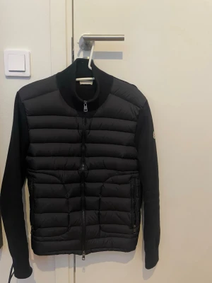 Moncler cardigan - Säljer en svart pufferjacka från Moncler med quiltad front, ribbade muddar och tvåvägsdragkedja. Jackan har Moncler-logga på ärmen och sidofickor med dragkedja. Materialet är en mix av dun och mjukt tyg på ärmar och rygg. Perfekt för kalla dagar.