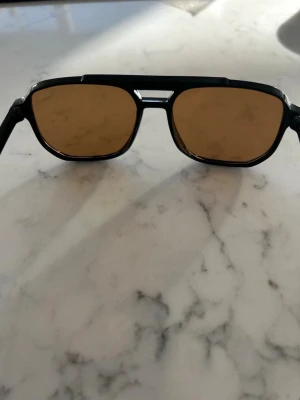 Svarta solglasögon med bruna glas - Snygga solglasögon med svart, kantig båge och bruna glas. Klassisk design med markerad näsbrygga och raka skalmar. Perfekta för soliga dagar och ger en cool vibe till din outfit.