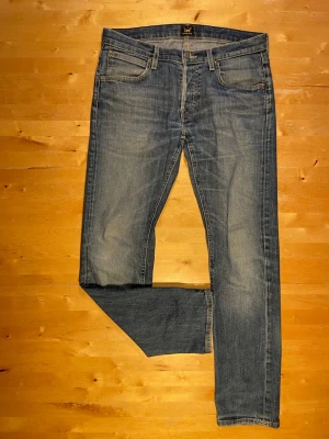 Blå raka jeans från Lee - Säljer ett par klassiska blå jeans från Lee med rak passform och fem fickor. Jeansen har snygga slitningar och en tidlös look med Lee-lapp bak i midjan. Perfekta för dig som gillar en enkel och stilren denimstil.