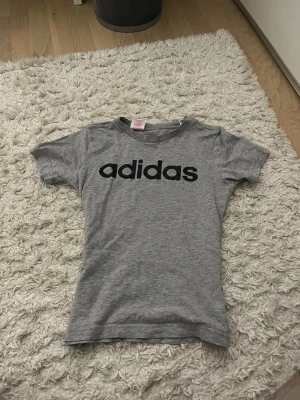 Grå adidas t-shirt med logga - Snygg grå t-shirt från adidas med klassisk svart logga tryckt över bröstet. T-shirten har rund halsringning och korta ärmar. Perfekt för en sportig och avslappnad stil. Tillverkad i mjukt bomullsmaterial.