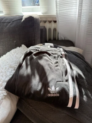Svarta Adidas mjukisbyxor med vita ränder - Säljer ett par svarta mjukisbyxor från Adidas med klassiska vita ränder längs benen och broderad logga på vänster sida. Byxorna har resår i midjan och sidofickor. Perfekta för chill eller träning.