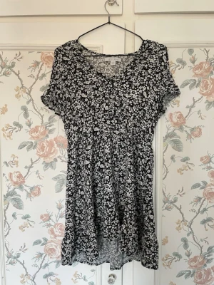Svart blommig kortklänning med volang - Supersöt svart kortklänning med vitt blommönster och volangkant. Klänningen har v-ringning och korta ärmar, vilket ger en luftig och somrig känsla. Perfekt för dig som gillar en feminin och avslappnad stil. Skriv vid funderingar 🩷