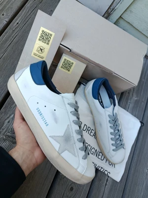 Golden Goose vita sneakers med blå detalj - Golden Goose sneakers i vitt läder med grå mockadetaljer och blå hälkappa. Klassisk stjärnlogga på sidan och grå skosnören. Snygg vintage-look och låg modell som passar till det mesta. Märket syns på hälen och sidan.
