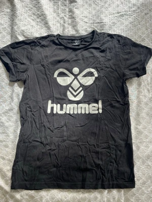 Svart Hummel t-shirt med logga - Snygg svart t-shirt från Hummel med stor vit logga framtill. Klassisk passform och rund halsringning. Perfekt till jeans eller shorts för en sportig look. Storlek 164 som passar till 14 år. 