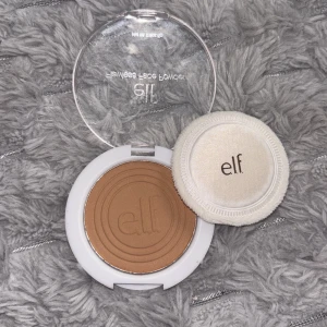 e.l.f. pressat puder foundation - Pressat puder foundation från e.l.f. i en rund vit dosa med transparent lock och medföljande applikator. Färgen är medium-brun och produkten innehåller salicylsyra 1%. Perfekt för att jämna ut hudtonen och motverka blemmor.
