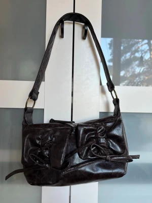 Handväska med rosetter - Snygg mörkbrun handväska. Väskan har två dekorativa rosetter på framsidan som även är fickor med magnetknapp. Perfekt storlek för det viktigaste och ger en cool vintagekänsla. Helt oanvänd.