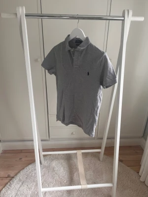 Grå piké från Ralph Lauren - Klassisk grå pikétröja från Ralph Lauren med korta ärmar och liten broderad logga på bröstet. Tröjan har krage och knappar vid halsen. Tillverkad i mjuk bomull som känns skön mot huden. Perfekt till jeans eller shorts för en clean look. Nypris ligger runt 1200kr.