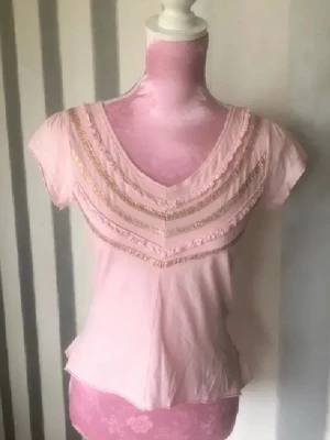 Ljusrosa topp med spets och volang - Supersöt ljusrosa topp med v-ringning, spetsdetaljer och volanger framtill. Korta ärmar och volangkant nedtill baktill ger en romantisk vibe. Perfekt för dig som gillar feminina detaljer och mjuka färger.