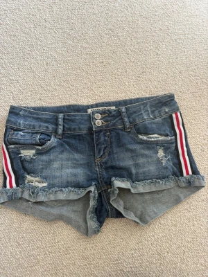 Blå jeansshorts  - Snygga blå jeansshorts med slitna detaljer och fransig kant. Shortsens sidor har röda och vita revärer som ger en sportig vibe. Två knappar framtill och klassiska fickor både fram och bak. Perfekta för sommaren!