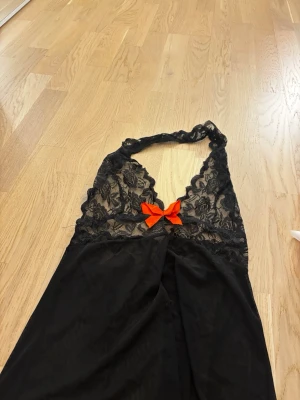 Svart spetsnattlinne med röd rosett - Snyggt svart nattlinne med halterneck och transparent spets upptill. Dekorerad med en röd rosett framtill för en extra detalj. Materialet är tunt och följsamt. Helt ny! 