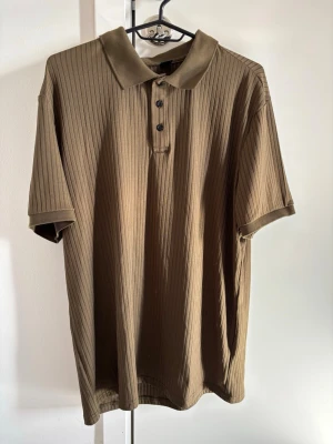 Ribbad brun kortärmad skjorta - Säljer en ribbad brun skjorta med korta ärmar och klassisk krage. Skjortan har knappar framtill och är tillverkad i ett mjukt material som känns skönt mot huden. Perfekt för dig som gillar en avslappnad men ändå stilren look.