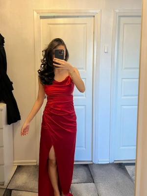 Röd sammetsklänning med slits - Elegant röd långklänning i satinmaterial med draperad överdel och smala axelband. Klänningen har en öppen rygg som ger en fin och feminin detalj samt en hög slits framtill som skapar en stilren silhuett.  Endast använd en gång till bal och i nyskick. Perfekt för fest, bal eller andra speciella tillfällen där du vill sticka ut.  Storlek 34. Pris kan diskuteras 