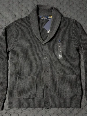 Grå cardigan från Polo Ralph Lauren - Mysig grå ribbstickad kofta från Polo Ralph Lauren med sjalkrage och knäppning framtill. Två stora fickor framtill och diskret blå logga broderad på bröstet. Perfekt för lager på lager och stilren look. STORLEK M, PASSAR S OCH M. HELT NY OANVÄND. 