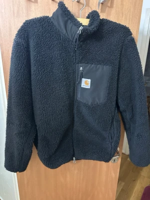 Svart teddyjacka från Carhartt - Mysig svart teddyjacka från Carhartt med hög krage och dragkedja framtill. Jackan har en bröstficka med dragkedja och Carhartt-logga, samt två sidofickor. Tillverkad i fluffigt teddy-material som håller dig varm under kyliga dagar.