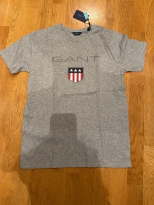 Grå GANT t-shirt med logga - Snygg grå t-shirt från GANT med klassisk logga och ett sköldmärke med stjärnor och ränder på bröstet. T-shirten har rund hals och är tillverkad i mjuk bomull, perfekt för en avslappnad och stilren look.