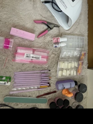 Stort nagelkit med UV-lampa och tillbehör - Säljer ett komplett nagelkit med UV-lampa, penslar i lila, nagelborstar, nageltippar, nagellim, fil, buffert, dotting tool, nageloljor, akrylpulver, geléer och remover. Perfekt för dig som vill göra egna naglar hemma. Flera färger på burkar och penslar, samt rosa och lila detaljer.