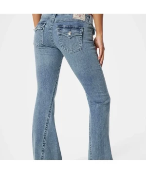 Blå bootcut jeans från True Religion - Säljer ett par klassiska blå bootcut jeans från True Religion med ikoniska fickdetaljer och kontrastsömmar. Jeansen har låg midja och utsvängda ben, samt dekorativa knappar på bakfickorna. Perfekta för en avslappnad och trendig look.