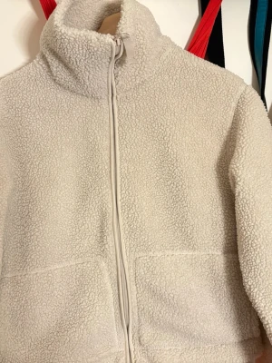 Beige teddyjacka från H&M - Mysig beige teddyjacka från H&M med hög krage och dragkedja framtill. Jackan har två stora fickor och är gjord i fluffigt teddy-material som håller dig varm. Perfekt för kyliga dagar och ger en trendig look till din outfit.