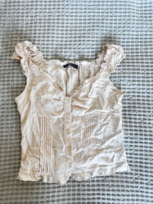 Ljusrosa blus med volang från Brandy Melville - Söt ljusrosa blus från Brandy Melville med knappar framtill, volangkant vid ärmhål och halsringning samt dekorativa veck på framsidan. Blusen är ärmlös och har en feminin, romantisk stil. Perfekt till jeans eller kjol.