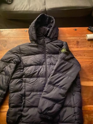 Svart dunjacka med huva Stone Island - Snygg svart dunjacka från Stone Island med huva och dragkedja framtill. Lite tunnare version av pufferjacka