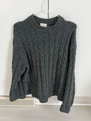Mörkgrå kabelstickad tröja H&M - Mysig mörkgrå kabelstickad tröja från H&M med rund halsringning och långa ärmar. Tröjan har ribbade muddar vid ärmslut och nederkant, perfekt för lager på lager och en chill look. Materialet är mjukt och värmande, passar till många olika outfits.