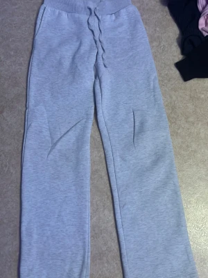 Ljusgrå mjukisbyxor med dragsko - Säljer ett par ljusgrå mjukisbyxor med bred resår i midjan och dragsko för skön passform. Byxorna har raka ben och sidofickor. Perfekta för chill dagar eller när du vill vara extra bekväm. Materialet känns mjukt och stretchigt.