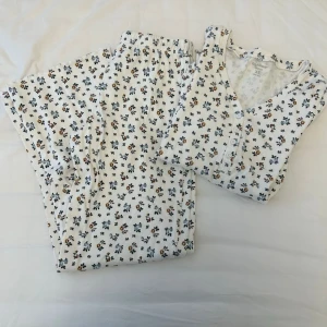 Blommig ribbad pyjamas från märket Mywear - Supersöt tvådelad pyjamas i ribbat material från märket Mywear, köpt på ICA Maxi i storlek XS. Setet är vitt med små blommor i blått och gult. Tröjan har V-ringning och knappar framtill, byxorna har resår i midjan med knytsnören. Armsluten och byxornas nederkant är vågiga. Aldrig använda. 
