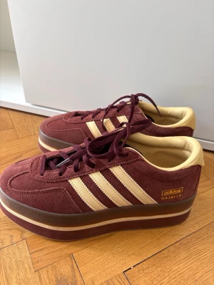 Adidas Gazelle vinröda platåsneakers - Säljer ett par Adidas Gazelle sneakers i vinrött mocka med beige detaljer och klassiska tre ränder. Skorna har en rejäl platåsula i vinrött och beige, samt matchande vinröda skosnören. Perfekta för dig som vill sticka ut med retrovibbar och cool siluett.