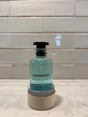 Louis Vuitton Imagination 100ml – NY/OANVÄND ✨ - Lyxig parfym från Louis Vuitton, Imagination, i en elegant glasflaska med ljusblå vätska och svart lock. Flaskan har en stilren, rund form och kommer med en vit cylinderformad förpackning med svart text. Perfekt för dig som gillar exklusiva dofter.