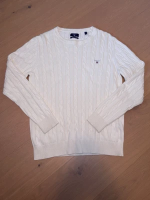 Vit kabelstickad tröja från GANT - Kabelstickad vit tröja från GANT med rund halsringning och broderad logotyp på bröstet. Tröjan har långa ärmar, ribbade muddar och nederkant. Klassisk och stilren design som passar till mycket.