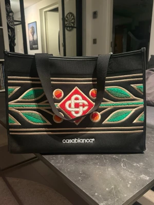 Casablanca Knitted shopping bag - Svart shoppingväska från Casablanca med broderat mönster i grönt, rött, orange och beige. Väskan har två handtag och tydlig logotyp framtill. Materialet är kraftig canvas och designen är rektangulär med färgglada detaljer.                  Väskan är helt ny med tag kvar!