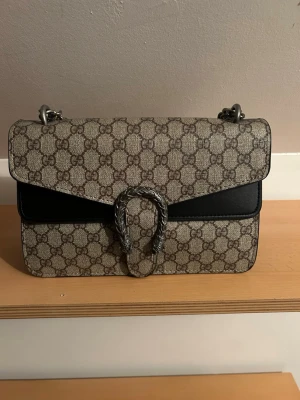 G*cci Dionysus monogram handväska - Säljer en G*cci handväska med klassiskt GG-monogram i beige och brunt, detaljer i svart läder och en snygg silverfärgad kedja. Väskan har ett dekorativt spänne framtill och flera fack inuti, inklusive ett med dragkedja. Perfekt för dig som gillar lyxig och tidlös design. Spännet på framsidan saknar en skruv som fester det, inget som syns vid användning. 