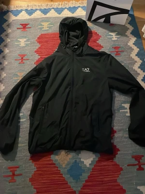 Svart jacka  från EA7 Emporio Armani - Ny pris 2000 Snygg svart jacka från EA7 Emporio Armani med dragkedja framtill och huva. Jackan har diskret logga på bröstet och är tillverkad i ett lätt, sportigt material med mesh foder på insidan. Perfekt för dig som gillar stilren och sportig look. 