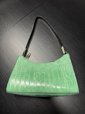 Grön axelväska i krokodilimitation - Snygg grön axelväska med croco-mönster och stilren form. Väskan har ett kort axelband i svart och stängs med dragkedja. Materialet är konstläder med glansig finish och väskan har guldfärgade metalldetaljer. Perfekt för dig som vill sticka ut med en trendig accessoar.