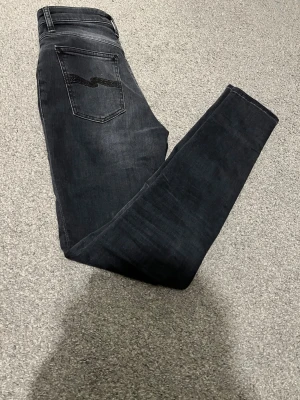 Svarta  jeans från Nudie Jeans - Säljer ett par svarta skinny jeans från Nudie Jeans med diskret slitning och klassisk femficksdesign. Jeansen har en svart broderad detalj på bakfickan och är tillverkade i stretchigt denim för en bekväm passform.
