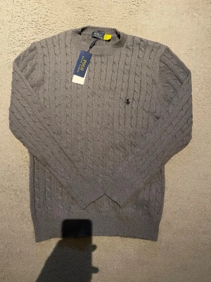 Grå kabelstickad tröja från Polo Ralph Lauren - Säljer en grå kabelstickad tröja från Polo Ralph Lauren med rund halsringning och diskret broderad logga på bröstet. Tröjan har klassisk passform och ribbade muddar vid ärmslut och nederkant. Perfekt för lager på lager under kyliga dagar.