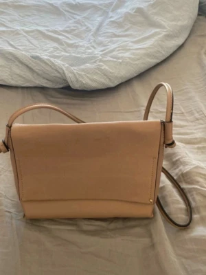 Beige axelväska i skinnimitation - Snygg beige axelväska med justerbar rem och magnetlås. Väskan har ett stilrent och minimalistiskt utseende med en stor ficka inuti. Perfekt för dig som gillar enkel och clean design. Materialet är skinnimitation och väskan har en mjuk struktur. Köpt på Åhléns. Finns ett litet märke ( se bild)