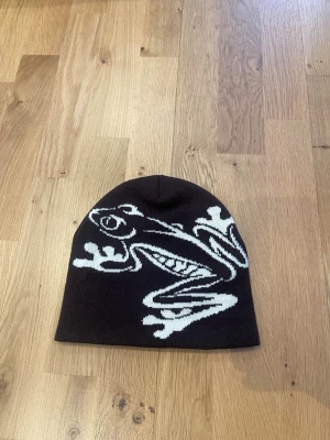Brun mössa med grodmotiv  - Snygg brun mössa med coolt vitt grodmotiv framtill. Mjuk och stretchig, sitter bra på huvudet och funkar både höst & vinter. 